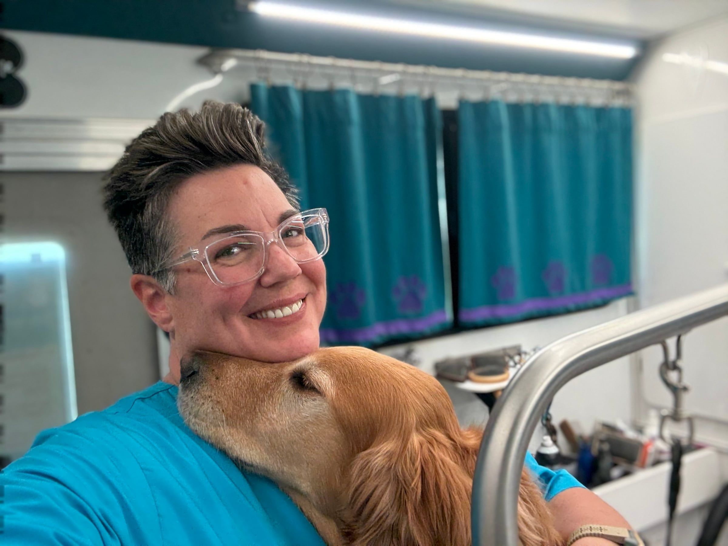 Amanda smiling as a golden retriever nuzzles her cheek inside the grooming trailer